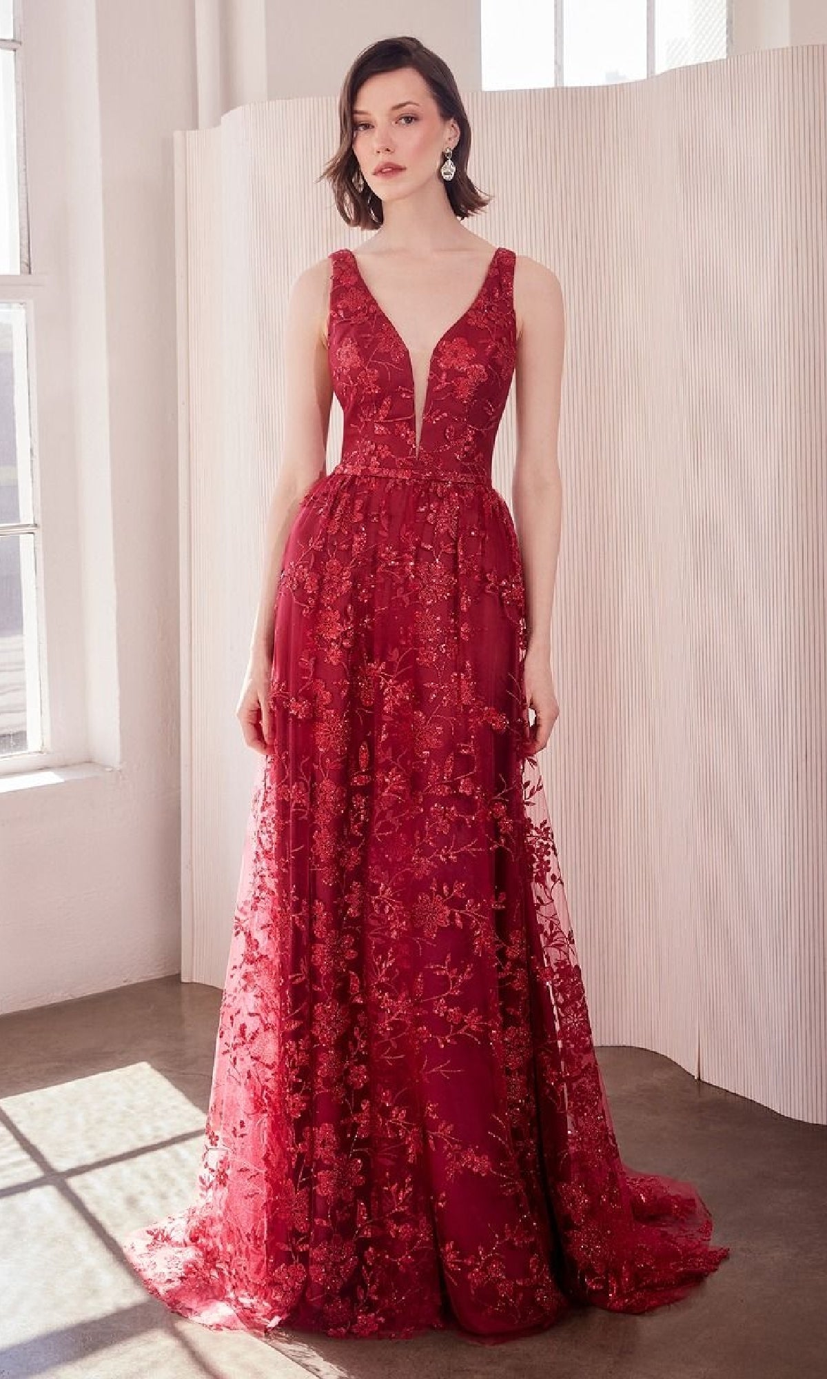 A woman stands indoors wearing the Amoris T606, an embroidered-lace long prom dress featuring a sleeveless, deep V-neck design with floral embroidery and a sheer overlay.
